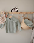 A wooden wall peg holds mushie Muslin Bibs, teething toys, a black-and-white baby photo, a mint green knit sweater, a woven straw bag with matching knit pants, and a beige robe against a light gray wall.