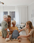 A woman and three kids gather around a cozy living room table, packing a light blue backpack with toys, baby accessories, and the mushie Silicone Pacifier Clip | Luna. The youngest child, wearing a knitted hat, stands on the table.