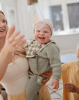 A smiling woman holds a laughing baby in green, chewing on the mushie Silicone Pacifier Clip | Hera, while a young boy in a yellow shirt stands beside them in a bright, cozy room.