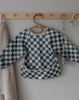 A blue and white checkered long-sleeve bib hangs on a wooden peg rack beside a beige textured towel and a chain featuring FRIGG Lucky Symmetrical Silicone Pacifiers (0-6 Months) for baby comfort and safety.