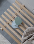 The mushie Cradle Cap Brushes, featuring gentle silicone bristles and a white holder, rest on a light wooden slatted bench beside a textured beige towel in a bathroom setting.