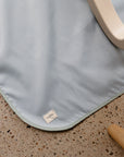 A light blue, water-resistant Mushie Splat Mat with rounded corners and a small MINIMAT tag rests on a terrazzo floor near light wood furniture—ideal as a high chair mat or splat mat.