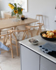A modern kitchen with a bowl of apples, a Mushie Baby Food Freezer Tray on the countertop, a wooden dining table with chairs, sunflowers in a vase, and soft natural light.