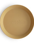 A round, tan mushie Round Dinnerware Plate made of non-toxic plastic features the “mushie” logo in the center and is photographed on a white background—ideal for everyday use.