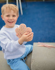Smiling boy holding a mushie Silicone Sensory Toy while sitting on a concrete bench outdoors.
