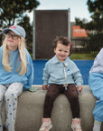 Three young children sit on a concrete bench outdoors. One child uses the FRIGG Daisy Night Silicone Pacifier from FRIGG, looking relaxed and happy with friends. A playground and trees can be seen in the background.