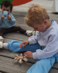 A young boy in a light blue outfit plays with the mushie Water Wheel Bath Toy on wooden boards outdoors, while a toddler behind him in casual clothes drinks from a cup and watches.