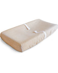 The mushie Extra Soft Muslin Changing Pad Cover, in beige and white stripes with safety straps, offers a cozy spot for your baby.