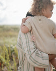 Outdoors, an adult in stripes carries a curly-haired toddler in a beige romper. The toddler holds a Mushie FRIGG Rope Natural Rubber Pacifier, clipped to their outfit, with greenery and sunlight in the background.