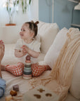 A young child sits on a white couch, smiling beside an adult. Nearby are colorful stacking toys and a shape sorter. A mushie Stretchy Swaddle is draped in the softly lit room, with a plant and other blankets in the background.