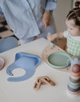 A young child in a green checkered outfit sits at a white table with colorful Mushie Classic Silicone Suction Plates and a blue bib, next to a toy chain and stacked ring toy, while an adult stands nearby.