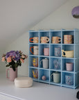 A blue cubby shelf with compartments displays colorful mugs and cups on a white countertop. Nearby are a bouquet of flowers in a pink vase, a mushie Lunchbox, and a coffee machine under a sloped ceiling with a purple wall lamp.