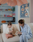 A woman and young child sit on a white couch in a colorful living room, using the mushie Shape Sorting Box for hands-on learning. Shelves with books, decor, and floral artwork adorn the cozy background.