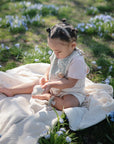 A toddler with braided hair sits on a blanket in the grass, surrounded by purple flowers and holding a FRIGG Butterfly Anatomical Silicone Pacifier (6-18 Months) from the 2-Pack, enjoying a sunny outdoor day.