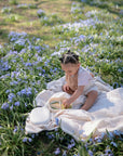 A young child, wearing a mushie Muslin Bib made of soft organic cotton, sits on a blanket among purple flowers, calmly exploring toys or snacks in their lap under the bright sun.