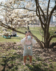 A toddler in a flower-embroidered outfit stands barefoot on grass, sipping from a cup beneath blossoms. A mushie Muslin Bib drapes softly around the child’s neck, with spring blooms and relaxed picnickers visible in the background.