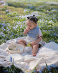 A toddler with styled hair sits on a picnic blanket outdoors among purple wildflowers, enjoying snacks from a mushie Lunchbox with built-in compartments. Sunlight filters through the grassy field.