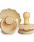 Two FRIGG Daisy Natural Rubber Pacifiers in Pale Daffodil are shown—one upright to display its latex nipple, the other flat with its scalloped shield and handle visible. This is the Limited Edition 2-Pack.