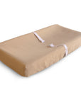 The mushie Extra Soft Muslin Changing Pad Cover in tan features a contoured design with a white safety strap across the middle, shown on a white background.