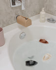 A white bathtub filled with water has several colorful toy boats floating in it. A pink cup, mushie Fragrance Free Baby Shampoo & Body Wash, and a bottle of soap rest on the tub’s edge, while a beige rainbow-shaped spout cover tops the faucet.