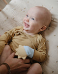 A smiling baby in a mustard ribbed onesie lies on a bee-print blanket, holding the mushie Sun Teething Mitten while an adult’s hands support them.