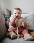 A smiling baby wearing a pink outfit and beige bib sits on a couch, holding the mushie Lamb Figurine Teether, surrounded by white and gray pillows.