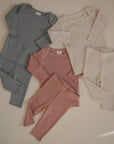 Three mushie Two Piece Sets for babies in blue, pink, and cream are laid flat on a neutral background. Each set includes a matching long sleeve shirt and pants, slightly overlapping for a coordinated display.