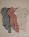 Three mushie Two Piece Sets in blue-gray, dusty rose, and cream are laid flat side by side, each featuring a long sleeve shirt and pants on a neutral background.