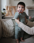 A baby wearing the mushie Two Piece Set stands on a couch, supported by an adult’s hands, chewing on fabric and gazing at the camera in a cozy, softly lit living room.
