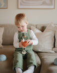 A blonde-haired child in green overalls and a white shirt sits on a beige couch holding a toy pumpkin, surrounded by light pillows, blue geometric toys, and FRIGG Little Viking Silicone 2-Pack pacifiers from FRIGG.