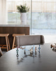 On a wooden table with modern chairs, mushie Daisy Silicone Suction Builders are arranged in pastel clusters. In the softly lit background, see a kitchen island with stools and a large vase with greenery.