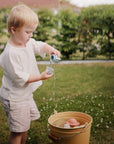A young child stands on grass, pouring water into a yellow bucket filled with mushie's Space Mold Free Bath Play Set, with toy boats and bath toys, greenery, and a building in the background.