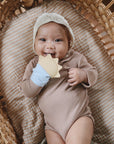 A baby in a brown outfit and bonnet lies in a wicker basket, smiling and holding the mushie Sun Teething Mitten, with the light blue mitten on one hand.
