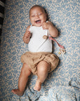A smiling baby lies on a blue and white floral mushie Organic Cotton Muslin Swaddle Blanket, wearing a white tank top and beige shorts with a pacifier clipped to their outfit.