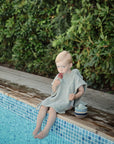 A young child sits poolside with their feet in the water, enjoying a rainbow popsicle made using the mushie Toddler Ice Pop Tray. Small bowls are beside them and green bushes form the background.
