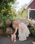 A young girl in a floral dress explores hands-on learning with the mushie Shape Sorting Box near wildflowers and a stone wall, outside a red barn on a sunny day.