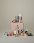 The mushie Shape Sorting Box features pastel geometric and heart-shaped pieces arranged around a pink box, providing hands-on learning and helping kids develop fine motor skills.