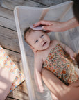 A smiling baby in a floral outfit lies in a clear tub during bath time as an adult gently uses the mushie Cradle Cap Brushes to brush their hair. The tub sits on a wooden floor beside a patterned blanket.
