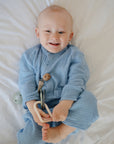 A smiling baby in blue pajamas lies on a white bed, holding the mushie Key Toy—a non-toxic teething toy—and looking up.