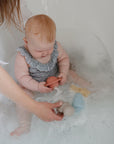 A baby in a blue patterned romper sits in a bubble-filled tub, playing with mushie’s Space Mold Free Bath Play Set. An adult hand offers another silicone toy from the set for more bathtime fun.