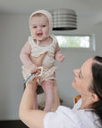 A woman with dark hair smiles up at a baby in a bonnet and light clothes, who is holding the mushie Key Toy (a non-toxic plastic toy), in a bright, modern room.