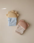 Two mushie Sun Teething Mittens are shown on a beige background: one light blue and sun-shaped, the other light pink with bear ears. Both silicone mittens have textured surfaces to soothe baby's gums.