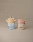 Two mushie Sun Teething Mittens stand upright on a neutral background: the left is blue with a yellow sun top, and the right is beige with a pink bear-ear and rainbow-shaped teether top.
