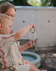 A young child sits on an adult’s lap outdoors, playing with the mushie Key Toy—linked, non-toxic plastic rings designed for teething. In the background, a small inflatable pool sparkles on the sunny patio.