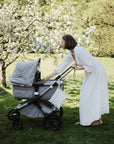 A woman in a long white dress leans over a gray stroller in a garden, reaching into the diaper bag storage for her mushie Water Resistant Wet Bag as she interacts with her baby.