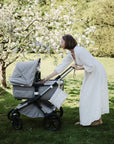 A woman in a long white dress leans over a gray stroller in a garden, reaching into the diaper bag storage for her mushie Water Resistant Wet Bag as she interacts with her baby.