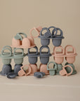 A collection of mushie silicone pacifiers, baby teethers, and the Toddler Ice Pop Tray in pastel pink, gray, and green are stacked neatly against a plain beige background.