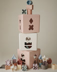 Three mushie Shape Sorting Boxes in pastel colors are stacked, each with cut-out shapes. Colorful geometric pieces scattered around inspire hands-on learning and fine motor skill development against a plain, neutral background.