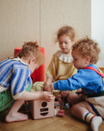 Three young children sit on a wooden floor, playing with the mushie Shape Sorting Box. Focused and engaged, they build fine motor skills as they explore the colorful blocks in a cozy indoor setting.