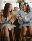 Two young girls sit on stools at a cozy kitchen counter, smiling and laughing. Both wear patterned outfits and boots, each holding mushie Square Dinnerware Plates, made from BPA-free plastic, on their laps.